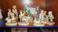Photo showing 9 ceramic figurines depicting children in various playful scenes including fishing, building a snowman, holding dolls, and sitting with animals.