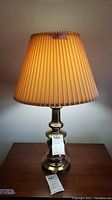 Full view of first brass toned table lamp with pleated beige shade standing on wooden surface, lit to show color and texture