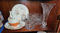Shelf display with plates, bowls, teacup and crystal vases showing detailed patterns and floral designs.