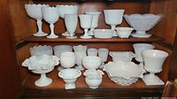 Wide shelf view showing multiple white opaque milk glass items including pedestal bowls, stemware, planters, and ruffled edge bowls.