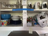 Shelf containing clear glasses, ceramic bowls, plates, mugs, glass chip and dip set, plastic bags with kitchenware
