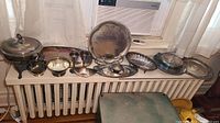 Full spread of silver-plated servingware pieces including chafing dish, platters, bowls, and buffet servers displayed on radiator and table.