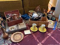 Wide view of lot showing Moroccan silverplate tea pot, vintage ashtrays, wooden box of assorted silverplate utensils, miniatures including dolls and animals, Bambi planter, two yellow ceramic cups on matching saucers, and a decorative tray.