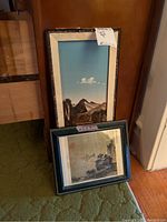 Two framed wall art pieces leaning against a wooden surface, one tall natural material mountain scene with blue sky and river, the other smaller Asian-inspired print.