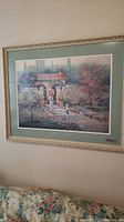 Framed wall art showing a house with tile roof surrounded by trees and garden pathway, pastel colors, ornate cream frame