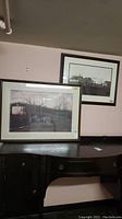 Two framed black and white wall art prints resting on a wooden table with a pink wall background