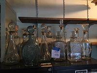 Group photo showing 13 assorted pressed glass decanters of various shapes and sizes arranged on a shelf, all with glass stoppers.