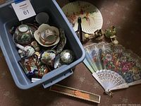 Blue storage bin filled with assorted floral tea cups, saucers, small ceramic items, and rickshaw figurines. Outside the bin are decorative folding fans with floral and landscape artwork on fabric and lace.
