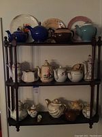 Front view of wooden display shelf holding multiple teapots on three levels, showing variety of colors and styles.