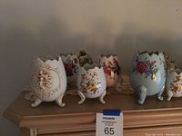 Multiple porcelain egg vases with floral designs, standing on three legs with scalloped tops, arranged on a wooden surface with a lace doily underneath.