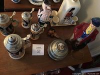 Top view of multiple German beer steins and ceramic decanters including one man figure in red coat and one in plaid, on wooden table surface.