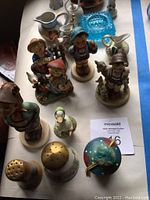 Tabletop with three Hummel figurines in traditional clothing, two salt and pepper shakers including mushroom and floral styles, small ceramic duck, blue glass ashtray, and ceramic miniature pitchers.