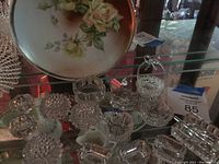 Multiple clear glass salt dishes, a floral round plate with roses, and part of the glass peacock dish.