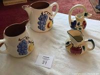 Four pitchers on white cloth: two larger Stangl pitchers with grape and pear motif, two smaller pitchers including one Purinton slipware.