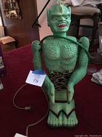 Green plastic humanoid Garloo figure seated on red carpet with power cord looped around torso