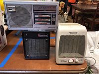 Full view of Panasonic radio on back with large speaker grille and controls, front view of black GoldStar heater, and off-white Pelonis ceramic heater.