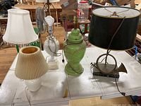 Five assorted lamps placed on a white table; two lamps missing shades; includes green ceramic, white ceramic with fabric shade, clear glass base, and lamps with pleated shades.