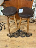 Two wooden barstools with round padded brown seats and three legs each, placed on a wooden floor. Two black metal scroll decorative pieces positioned in front of the stools.