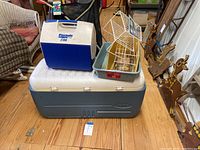 Photo showing the large Igloo cooler, small Playmate cooler, white wire kitchen storage rack, and a gray plastic bin with a red label all on a wooden floor.