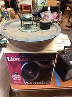 Photo showing Vornado Vortex electronic space heater box with the heater out of the box not visible, next to vintage bird fountain on table.