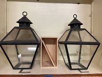 Two black metal and glass exterior lamp tops with a wooden box placed between them on a shelf.