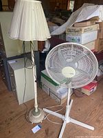 Floor lamp with pleated cream fabric shade positioned beside white Holmes floor fan against brown wooden floor and stacked boxes background.