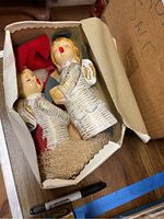Two angel figurines with painted faces and gold hair in a box containing red fabric and a decorative base.