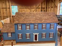 Front view of large two-story blue doll house with white window frames, burgundy shutters and front door, cedar-like shingled roof, mounted on a wooden base.