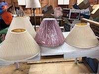 Five large lampshades including three off-white fabric and one pink fabric, displayed on a table.