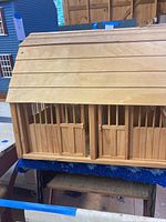 Wooden toy barn showing roof and sliding stable doors, placed over blue rug with wooden fence elements visible.