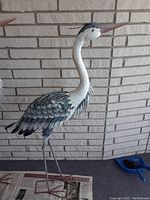 Side view of full metal crane statue showing long legs and detailed feather body against brick background.