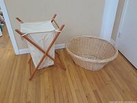 Photo showing the folding hamper with wooden frame and cloth bag alongside the oval wicker laundry basket on a hardwood floor with white walls.