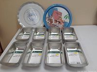 Photo showing eight stainless steel loaf pans with The Kitchen Company labels stating 'Loaf Pan Moule A Pain' and two metal trays, one commemorative and one decorative with animals.