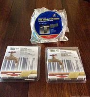 Three items on a wooden surface: two clear plastic packages containing the 9 ft and 6 ft heated freeze protection coils respectively, and a sealed roll of adhesive pipe insulation wrap.
