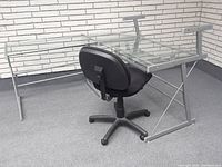 Full view of tempered glass office desk with metal frame and black fabric office chair on casters.