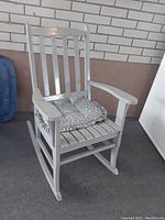 Front angle view of white wooden rocking chair with floral seat cushion against brick wall background.
