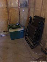 Black foldable anti gravity chair leaning against wall next to green Igloo cooler