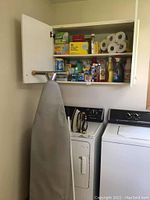Full view of wall cabinet above washer and dryer filled with cleaning supplies, paper towels, and cleaning boxes.