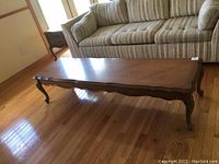 Full view of the wooden French provincial style coffee table placed on hardwood floor next to sofa showing curved edges and cabriole legs.