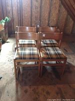 Four wooden dining chairs with plaid upholstered seats arranged together in dimly lit room.