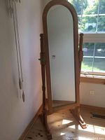 Full view of the full-length swivel cheval mirror with oval top and oak-finish wooden stand positioned near a window with light reflections visible.
