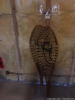 Photo showing a pair of 38 inch wooden snow shoes with woven webbing and leather foot bindings placed vertically against a wall.