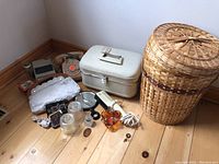 Wide view of all lot items on hardwood floor including rotary phone, luggage case, wicker basket, glass jars, magnifying glass and amber pitcher.