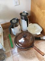 Full view of two CorningWare coffee pots, covered casserole dish, and Vision frying pan with lid on tiled floor.