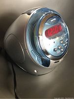 Side and front view showing the digital red LED clock display with control buttons and speaker side panel on the silver plastic body.