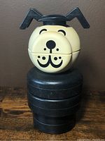 Front view of the vintage Fisher Price Lucky Dog stacking toy showing yellowish-beige head with black facial features and black ears on stacked black rings body.