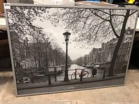 Full view of the large black and white cityscape photo framed with a visible red bicycle at the canal side, showing overall context and scale.