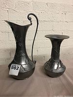 Pair of pewter ewer and vase placed on table against white brick wall, showing raised vine and leaf decoration