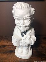 Full front view of the white plastic Colonel Sanders coin bank standing on wooden surface.