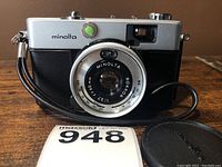 Front view of vintage Minolta Hi-Matic C 35mm film camera with wrist strap and lens cap placed nearby.